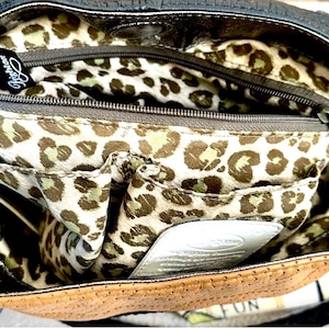May include: A handbag interior featuring a cream-colored lining with a brown and black leopard print pattern. The bag includes a zippered compartment with a black Brighton brand tag, an open pocket, and a white leather patch with embossed text.