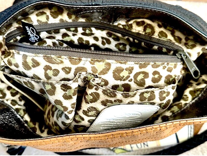 May include: A handbag interior featuring a cream-colored lining with a brown and black leopard print pattern. The bag includes a zippered compartment with a black Brighton brand tag, an open pocket, and a white leather patch with embossed text.