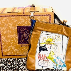 May include: A tan perforated leather shoulder bag featuring a graphic print of a person in a hat with the text DIVA, CHIC and FUN, GLAM GIRLS COUTURE PARIS, and VIP. The bag has a blue interior lining and a detachable strap with a metal clasp.