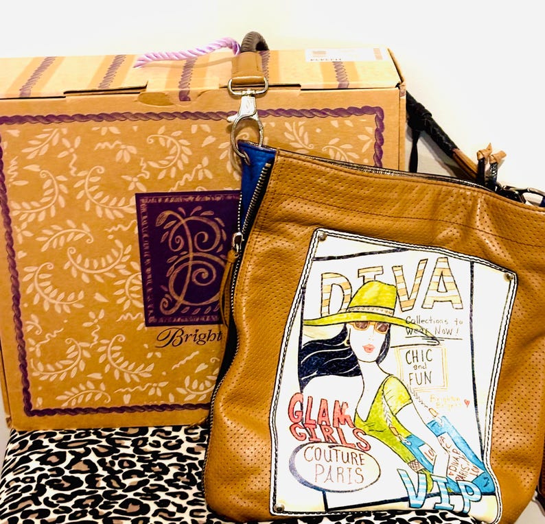 May include: A tan perforated leather shoulder bag featuring a graphic print of a person in a hat with the text DIVA, CHIC and FUN, GLAM GIRLS COUTURE PARIS, and VIP. The bag has a blue interior lining and a detachable strap with a metal clasp.