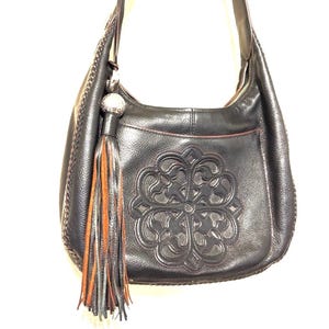 May include: Black leather shoulder bag with a decorative embossed floral design. The bag features a long strap, a tassel with orange and gray accents, and a zipper closure. The bag has a stitched trim.