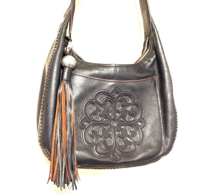 May include: Black leather shoulder bag with a decorative embossed floral design. The bag features a long strap, a tassel with orange and gray accents, and a zipper closure. The bag has a stitched trim.