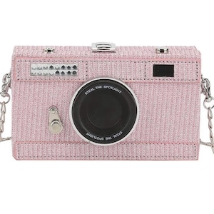 May include: A pink glitter camera-shaped crossbody bag with a silver chain strap and a black lens that says "Steal the Spotlight".