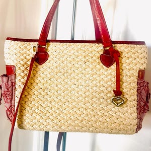 May include: A woven straw tote bag with red leather handles and trim. The bag features red heart-shaped accents and a gold heart charm. Red fabric pockets with floral patterns are on the sides.