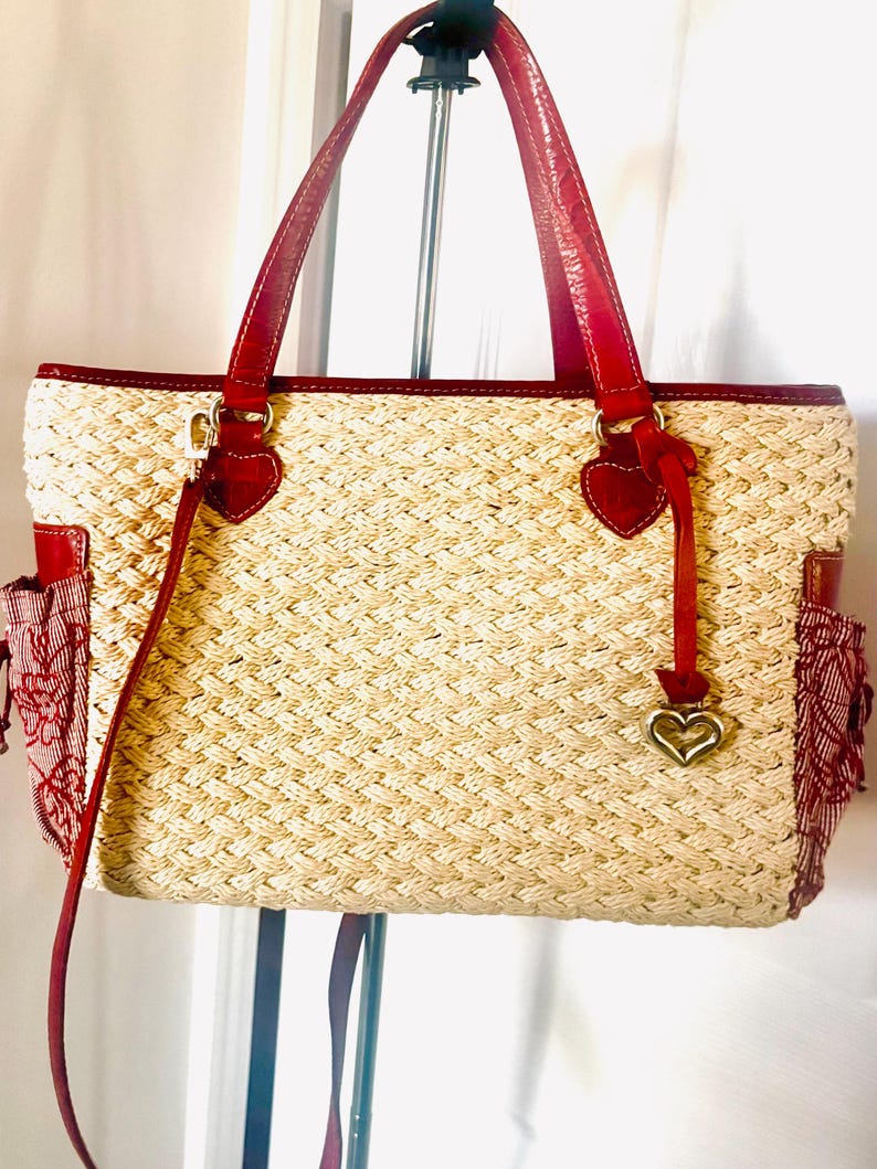 May include: A woven straw tote bag with red leather handles and trim. The bag features red heart-shaped accents and a gold heart charm. Red fabric pockets with floral patterns are on the sides.