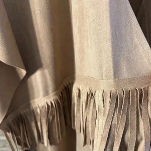 May include: Close-up of a tan garment with a fringed hem. The fabric appears to be suede or a suede-like material. The fringe is long and hangs down from the bottom edge of the garment, creating a textured look. The garment is draped.