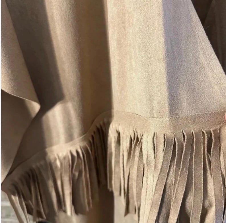 May include: Close-up of a tan garment with a fringed hem. The fabric appears to be suede or a suede-like material. The fringe is long and hangs down from the bottom edge of the garment, creating a textured look. The garment is draped.