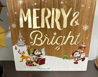 Könnte beinhalten: Ein Holzschild mit einem beleuchteten Design, das Mickey Mouse und Pluto in einer Winterszene zeigt. Das Schild trägt die Aufschrift "Merry & Bright" in goldenen Buchstaben.