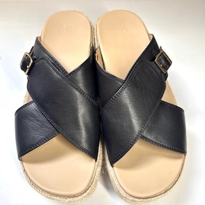May include: A pair of black leather sandals with a crisscross design. The sandals have a beige footbed and a woven jute sole. Each sandal features a gold-colored buckle. The sandals are designed for casual wear.