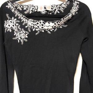 May include: A black long-sleeved sweater with white floral embroidery around the neckline and on the left shoulder. The embroidery features detailed flower and leaf patterns. The sweater is displayed on a black hanger.