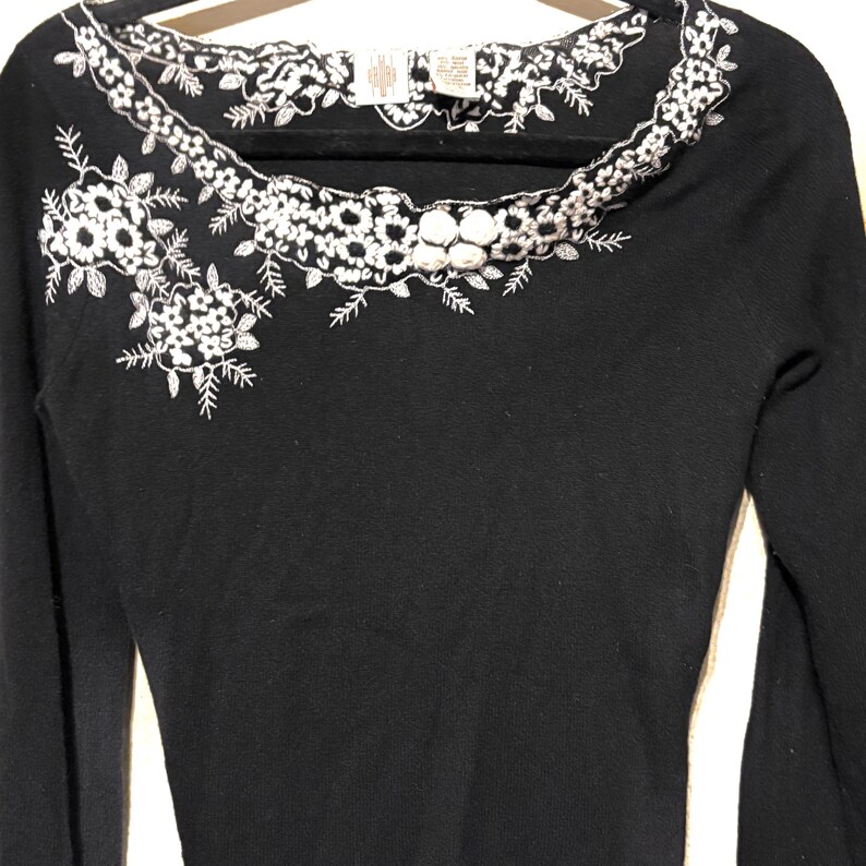May include: A black long-sleeved sweater with white floral embroidery around the neckline and on the left shoulder. The embroidery features detailed flower and leaf patterns. The sweater is displayed on a black hanger.