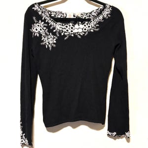 May include: Black long-sleeve sweater with white floral embroidery around the neckline and on the left shoulder. The cuffs also feature white floral detailing. The sweater is hanging on a black hanger against a white background.