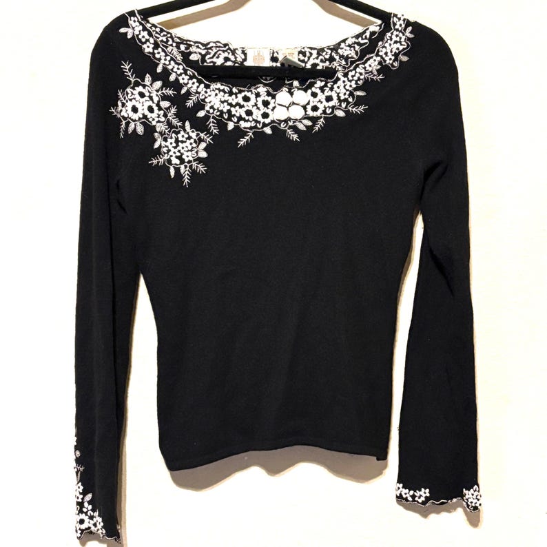 May include: Black long-sleeve sweater with white floral embroidery around the neckline and on the left shoulder. The cuffs also feature white floral detailing. The sweater is hanging on a black hanger against a white background.