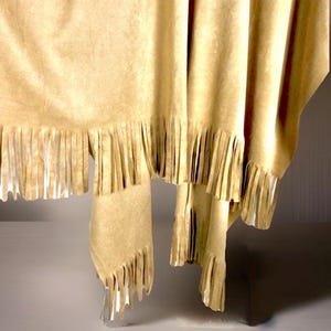 May include: A tan-coloured textile, possibly a shawl or throw, with a fringed edge. The fabric hangs in soft folds, and the fringe is evenly cut along the bottom. The material appears to be suede or a suede-like material, adding a tactile element.