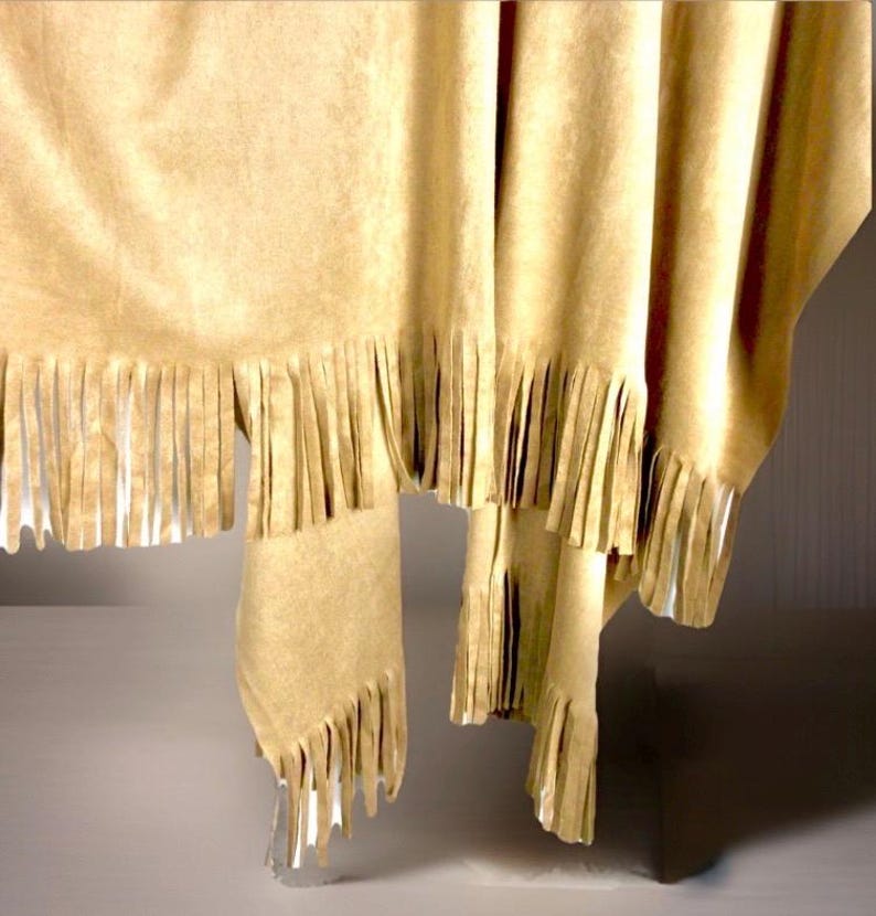 May include: A tan-coloured textile, possibly a shawl or throw, with a fringed edge. The fabric hangs in soft folds, and the fringe is evenly cut along the bottom. The material appears to be suede or a suede-like material, adding a tactile element.