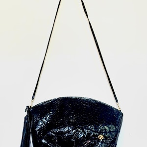 May include: A black crossbody bag with a glossy, textured finish resembling reptile skin. The bag features a curved top, a long thin shoulder strap, and a decorative tassel attached to the side zipper. A small gold-toned circular emblem is visible.