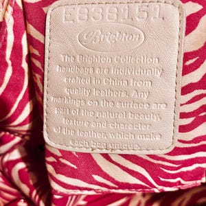 Pode incluir: Close-up de uma etiqueta de bolsa Brighton. A etiqueta &eacute; de cor bege claro com o nome da marca e texto. O fundo &eacute; um tecido com estampa de zebra vermelha e branca.