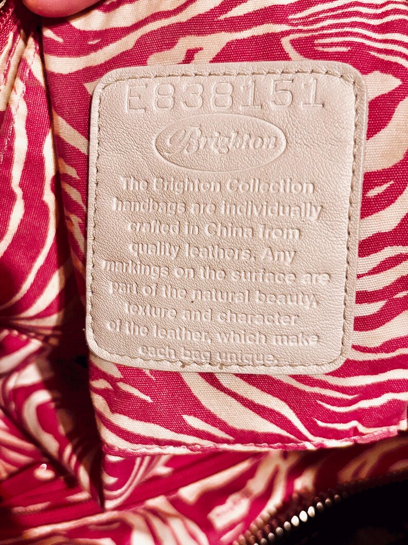 Pode incluir: Close-up de uma etiqueta de bolsa Brighton. A etiqueta &eacute; de cor bege claro com o nome da marca e texto. O fundo &eacute; um tecido com estampa de zebra vermelha e branca.