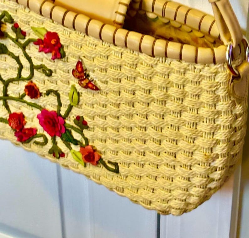 Puede incluir: Un bolso de paja tejido con un asa de cuero marr&oacute;n claro y un dise&ntilde;o floral. Las flores son rojas, rosas y verdes.