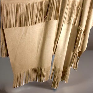 May include: A tan fringed poncho. The garment is made of a soft, flowing material with long fringe along the bottom and edges. The fabric has a subtle texture and drapes loosely, creating a relaxed look.