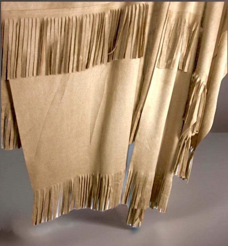 May include: A tan fringed poncho. The garment is made of a soft, flowing material with long fringe along the bottom and edges. The fabric has a subtle texture and drapes loosely, creating a relaxed look.