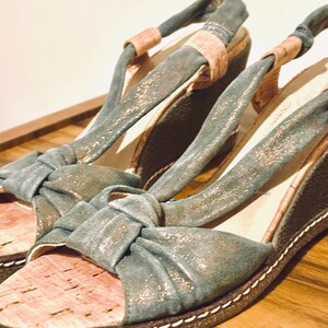 May include: A pair of teal wedge sandals with a cork footbed and knotted straps. The sandals have a metallic sheen and a white stitched border. The background is a wooden surface.