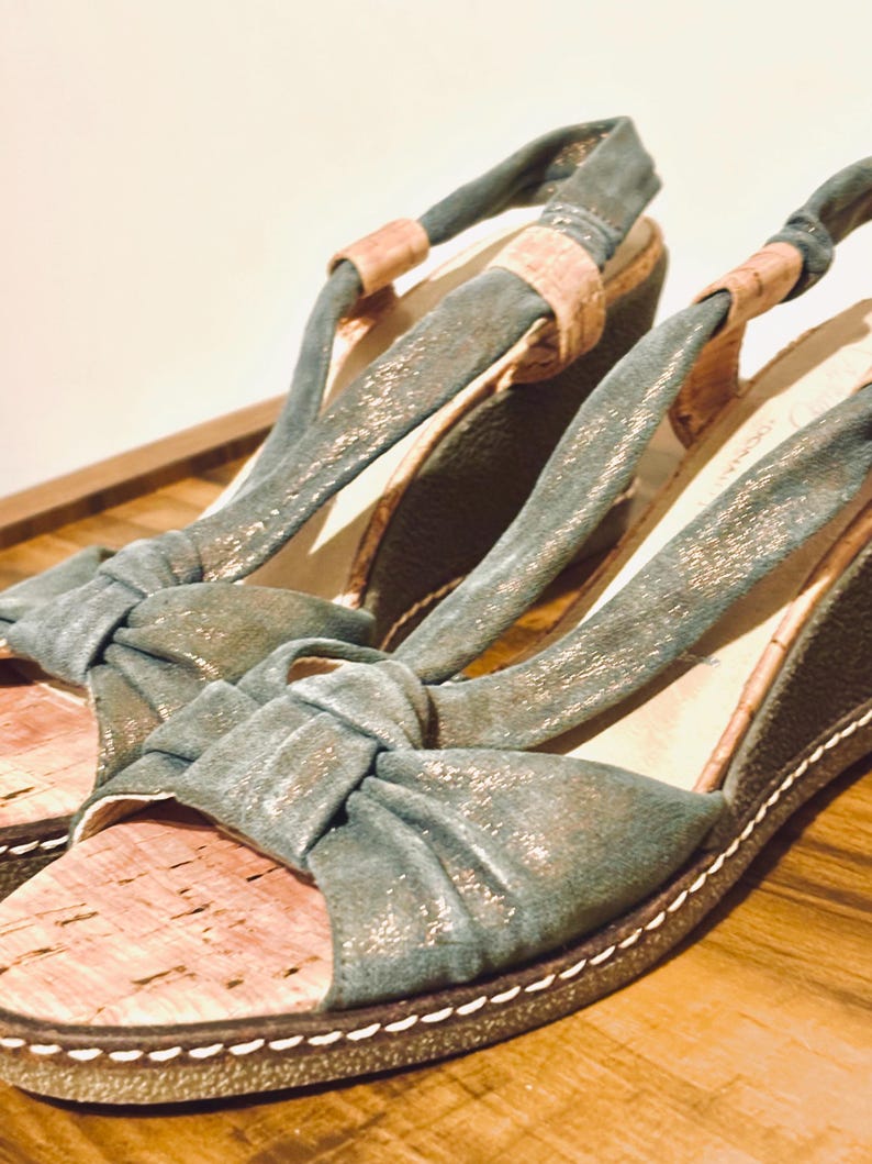 May include: A pair of teal wedge sandals with a cork footbed and knotted straps. The sandals have a metallic sheen and a white stitched border. The background is a wooden surface.