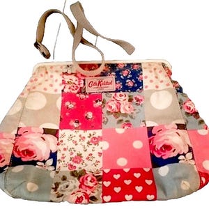 May include: A patchwork handbag with a floral and polka dot pattern. The bag features a variety of colors, including pink, red, blue, and gray. It has a gray handle and a clasp closure. The label reads "Cath Kidston".