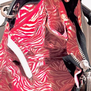 Pode incluir: Bolsa preta com forro de estampa de zebra vermelha e branca. A bolsa tem z&iacute;per e ferragens prateadas e uma al&ccedil;a branca. O interior possui v&aacute;rios bolsos. A bolsa est&aacute; aberta, revelando o design interior.