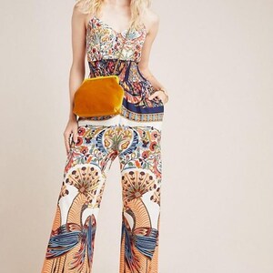 May include: A woman wearing a white and blue floral print jumpsuit with a yellow velvet clutch bag. The jumpsuit has a wide-leg silhouette and a fitted bodice. The woman is wearing her hair down and has a natural makeup look.