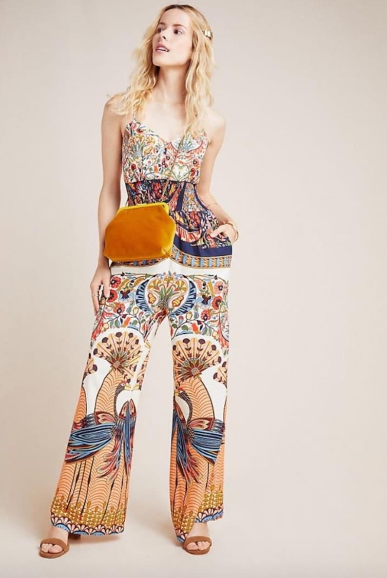May include: A woman wearing a white and blue floral print jumpsuit with a yellow velvet clutch bag. The jumpsuit has a wide-leg silhouette and a fitted bodice. The woman is wearing her hair down and has a natural makeup look.