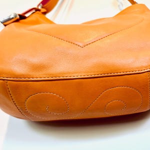 以下が含まれることがあります： オレンジ色の革製ハンドバッグのクローズアップ。バッグは曲線的な形状で、底にはステッチのデザインがあります。ストラップは上部にあり、シルバーのバックルが付いています。バッグは滑らかな革製です。