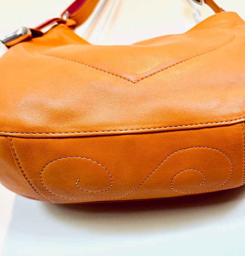 以下が含まれることがあります： オレンジ色の革製ハンドバッグのクローズアップ。バッグは曲線的な形状で、底にはステッチのデザインがあります。ストラップは上部にあり、シルバーのバックルが付いています。バッグは滑らかな革製です。