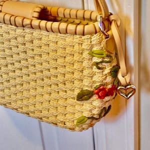 Puede incluir: Un bolso de paja tejido con ribete de cuero marr&oacute;n claro y un dije en forma de coraz&oacute;n. El bolso est&aacute; decorado con flores y hojas bordadas en tonos verdes, rojos y naranjas.