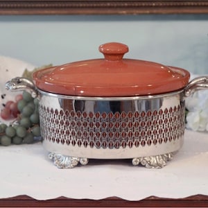 May include: A vintage terracotta-colored casserole dish with a silver-colored metal filigree base and lid. The dish has ornate handles and sits on decorative feet. The lid has a rounded knob. A classic piece for serving or display.