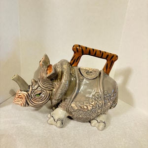 May include: A ceramic teapot shaped like a rhinoceros with a tiger-striped handle. The teapot is gray with brown and white details. The rhinoceros has a green eye and a textured surface.