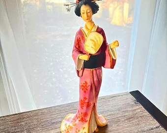 May include: A standing figurine of a person wearing a pink kimono with a red floral pattern, a black obi sash, and a gold-toned hair accessory with a butterfly charm. The figure holds a small, light-colored handheld fan in one hand.