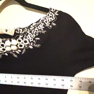 May include: Black garment with white floral embroidery along the neckline. The embroidery features small, round, and leaf-shaped details. A ruler is placed along the bottom edge, indicating measurements in inches.