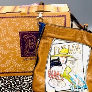 May include: A tan perforated leather shoulder bag featuring a graphic illustration of a person in a hat with the text DIVA, CHIC and FUN, GLAM GIRLS COUTURE PARIS, and VIP. The bag includes a blue interior lining and a metal clasp attachment.