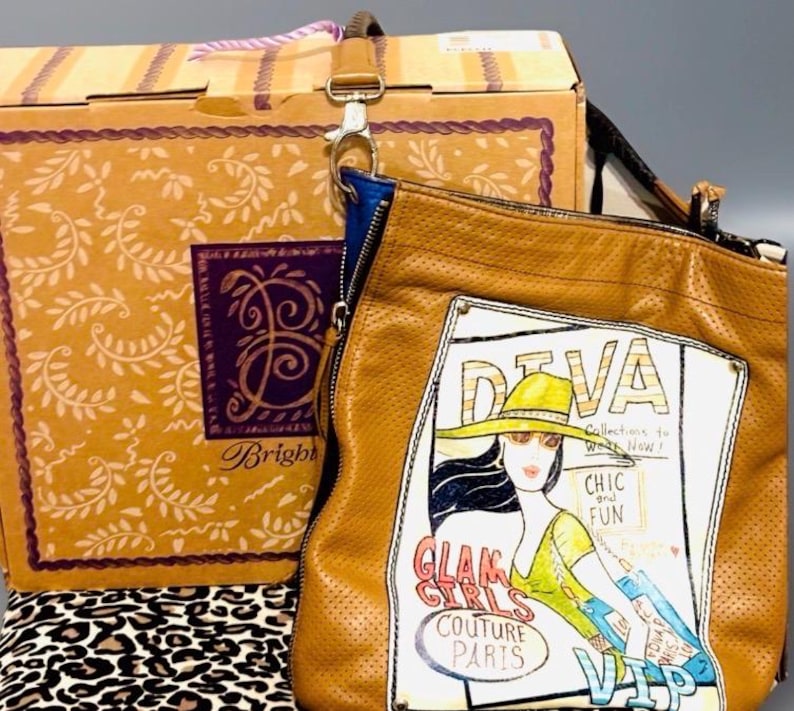 May include: A tan perforated leather shoulder bag featuring a graphic illustration of a person in a hat with the text DIVA, CHIC and FUN, GLAM GIRLS COUTURE PARIS, and VIP. The bag includes a blue interior lining and a metal clasp attachment.
