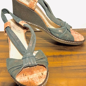 May include: A pair of wedge sandals with a cork footbed and a dark teal fabric upper. The sandals have a bow detail on the toe and a slingback strap. The wedge heel is approximately 3 inches high.