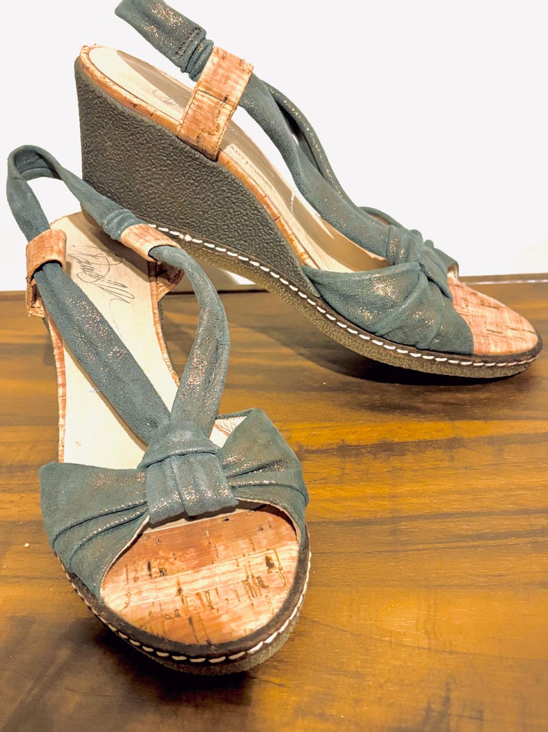 May include: A pair of wedge sandals with a cork footbed and a dark teal fabric upper. The sandals have a bow detail on the toe and a slingback strap. The wedge heel is approximately 3 inches high.