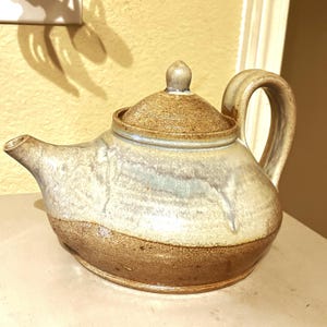 May include: A brown and white ceramic teapot with a lid. The teapot has a rounded body and a spout on one side. The handle is on the opposite side of the spout. The teapot has a brown glaze on the bottom and a white glaze on the top.
