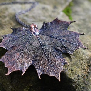 May include: A copper maple leaf pendant necklace with a detailed vein pattern. The pendant is hanging on a silver chain and resting on a rock.