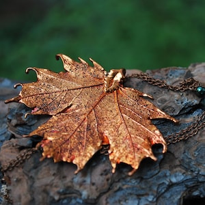 May include: A copper leaf pendant necklace with a chain and two dark green beads. The necklace is resting on a piece of tree bark.