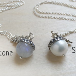 Op de afbeelding: Twee zilveren kettingen met eikelvormige hangers. De hanger aan de linker ketting heeft een maansteen, terwijl de hanger aan de rechter ketting een witte parel van schelp heeft.