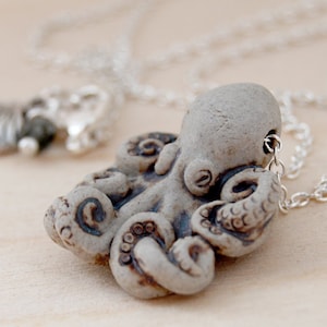 Op de afbeelding: Een zilveren ketting met een keramische octopus hanger. De octopus is lichtgrijs met bruine details. De hanger is met een zilveren lus aan de ketting bevestigd.