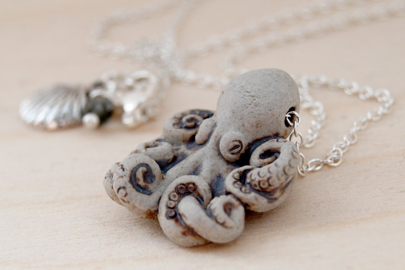 Op de afbeelding: Een zilveren ketting met een keramische octopus hanger. De octopus is lichtgrijs met bruine details. De hanger is met een zilveren lus aan de ketting bevestigd.