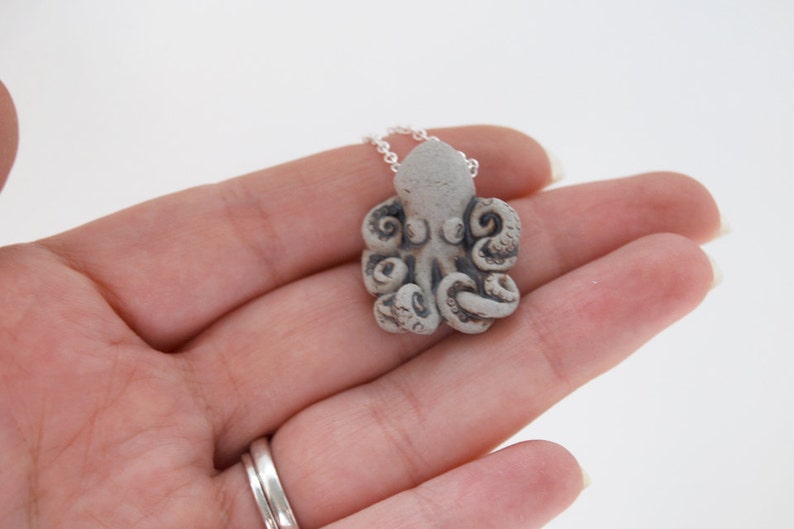 Op de afbeelding: Een zilveren ketting met een grijze keramische octopus hanger. De octopus heeft gedetailleerde tentakels en een getextureerd oppervlak.