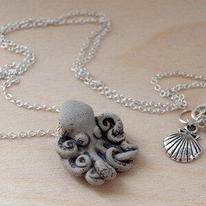 Op de afbeelding: Een zilveren ketting met een grijze keramische octopus hanger en een zilveren hanger van een schelp met een grijze edelsteen kraal.